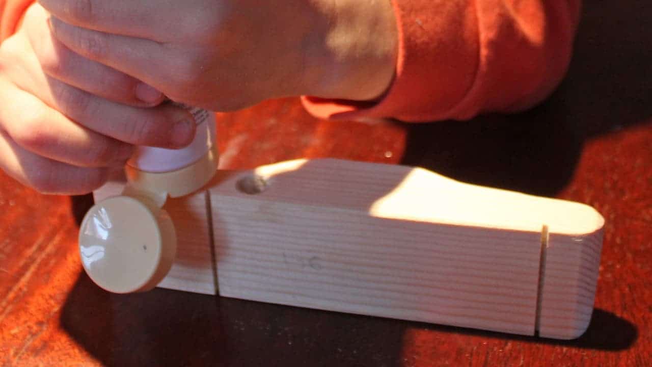 Hands squeezing wood filler from a tube onto a pinewood derby car body, smooth light-colored wood with drilled holes and marker lines