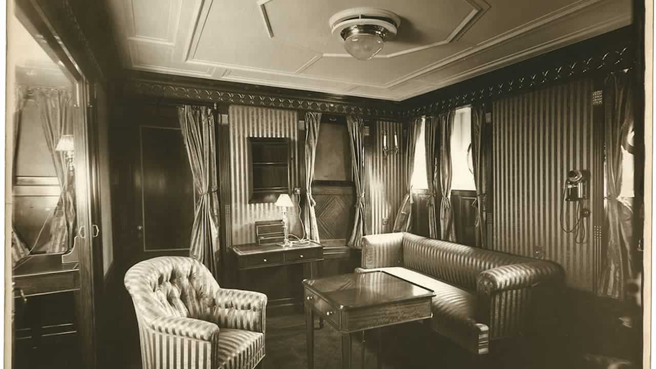 Vintage black and white photograph of elegant parlor room with striped upholstered furniture, ornate ceiling, curtained windows, decorative molding