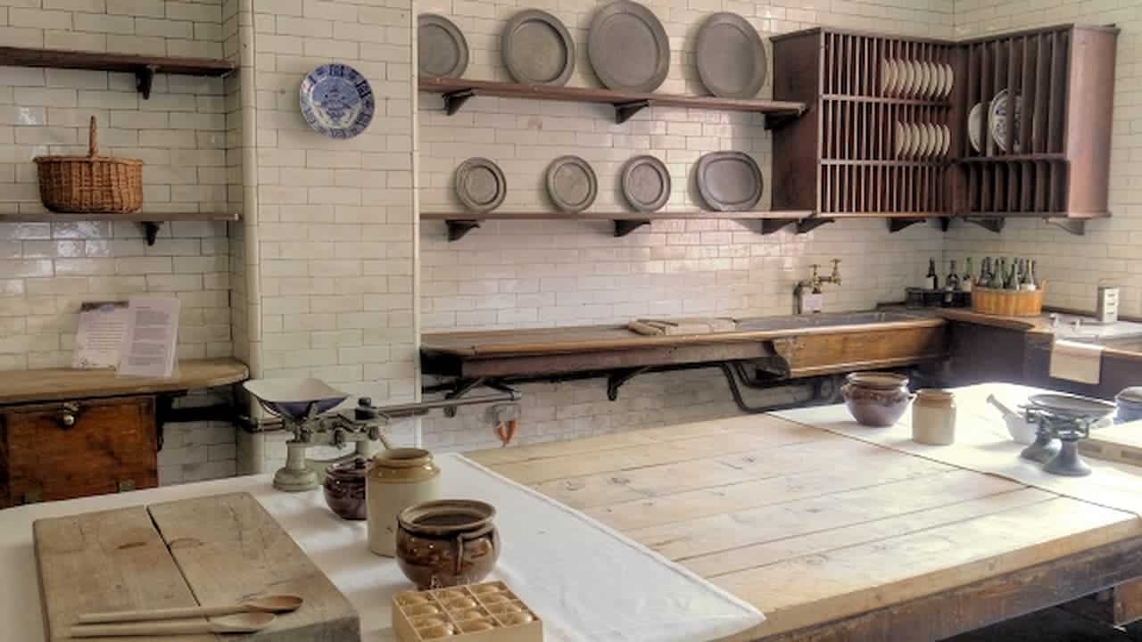 Historic scullery with white subway tile walls, wooden shelving displaying pewter plates, large work table, traditional kitchen workspace