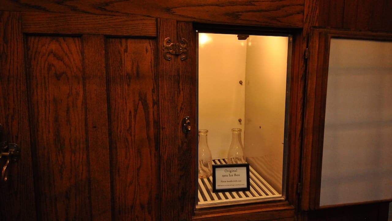 Vintage wooden icebox with open door revealing glass milk bottles and sign reading "Original 1902 Ice Box" inside