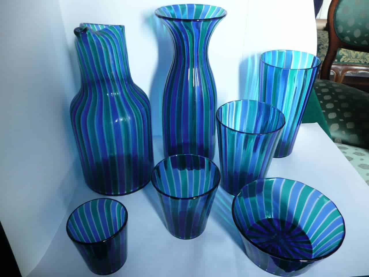 Set of striped Murano glassware in shades of blue, teal, and purple, including two pitchers, four tumblers, and a bowl, displayed against a white background
