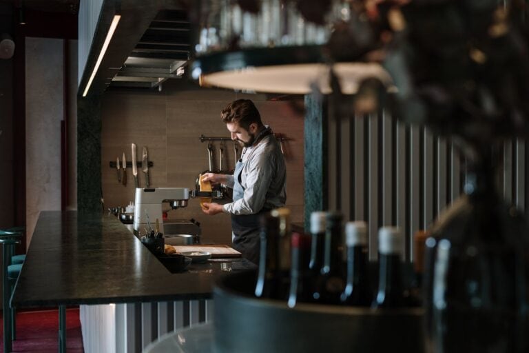 Person in white shirt working at a bar or restaurant counter with bottles lined up in the background, dim atmospheric lighting