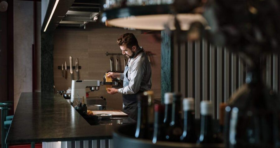 Person in white shirt working at a bar or restaurant counter with bottles lined up in the background, dim atmospheric lighting