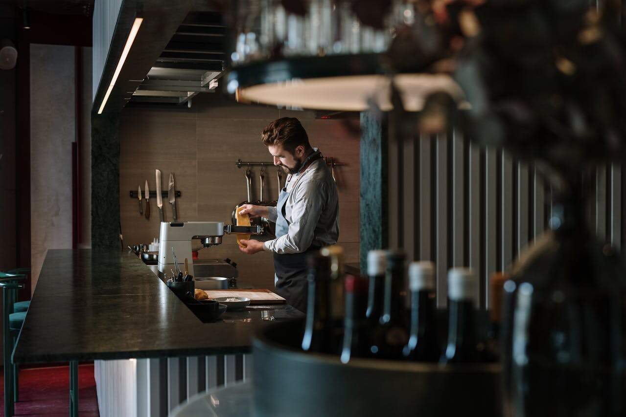 Person in white shirt working at a bar or restaurant counter with bottles lined up in the background, dim atmospheric lighting