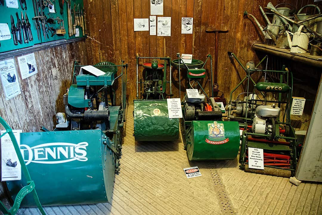 Museum display of vintage lawn mowers including Dennis, Ransomes, and Atco models in glass cases with wooden walls.