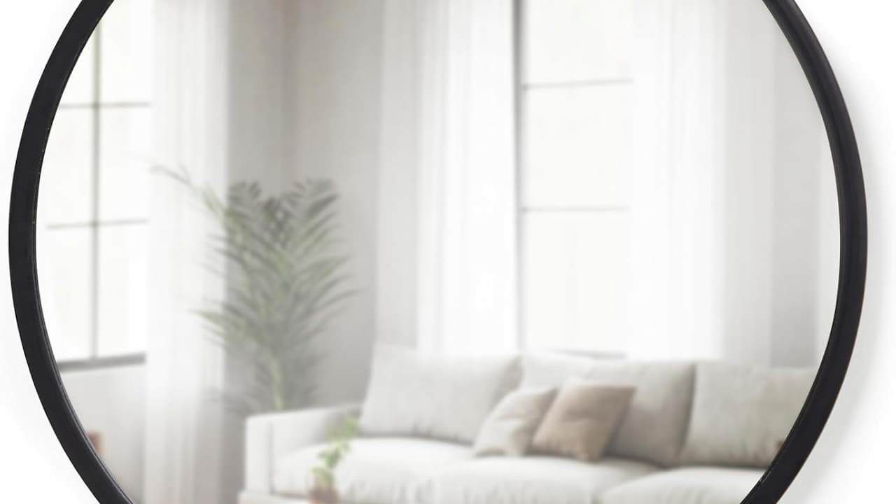 Round black-framed mirror reflecting bright living room with white sofa, pillows, and potted plant near window