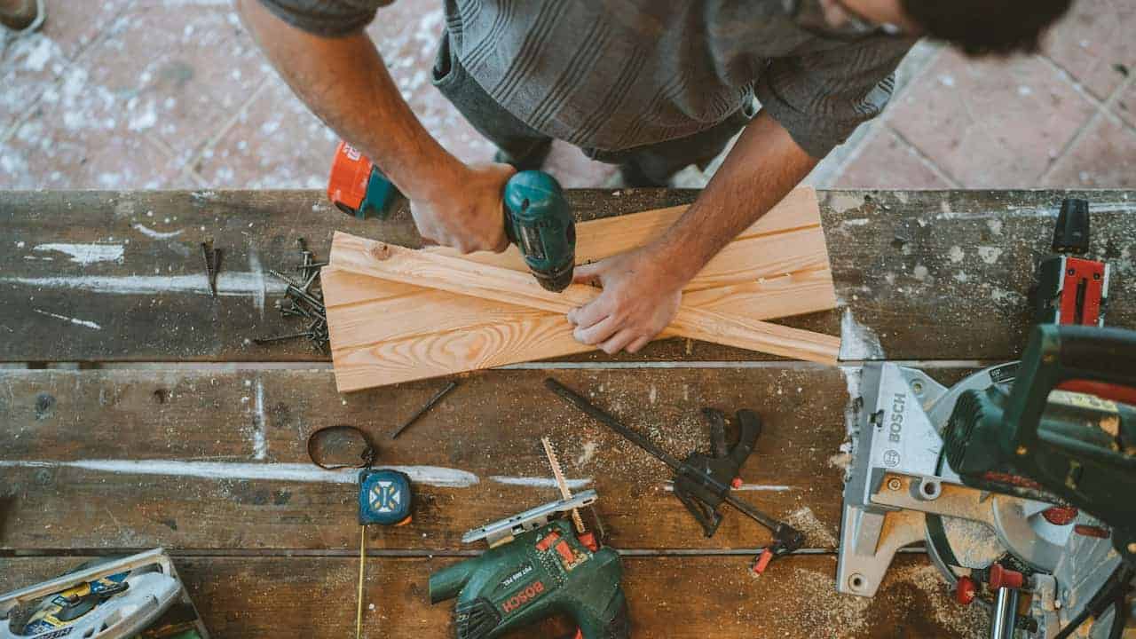 Man using a power drill on intersecting wooden planks, aligning and securing the wood with one hand while drilling with the other