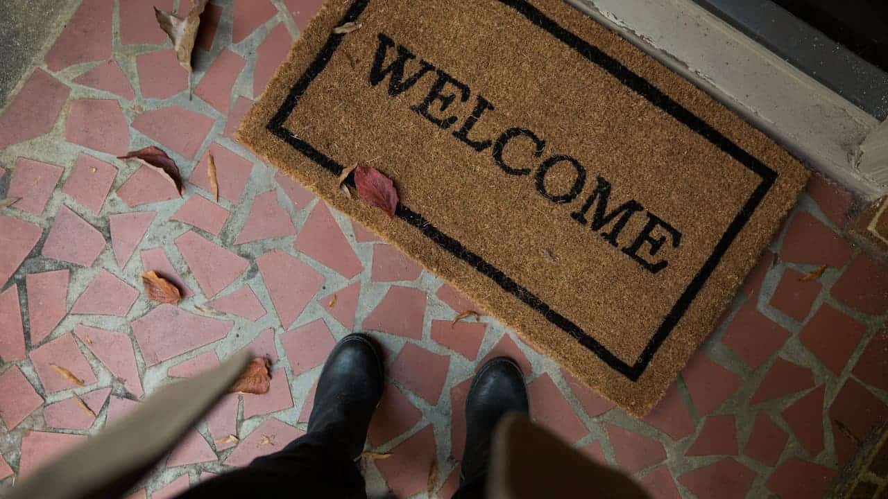Brown coir welcome mat with black border on broken red brick patio, with person's black shoes and dog paw visible