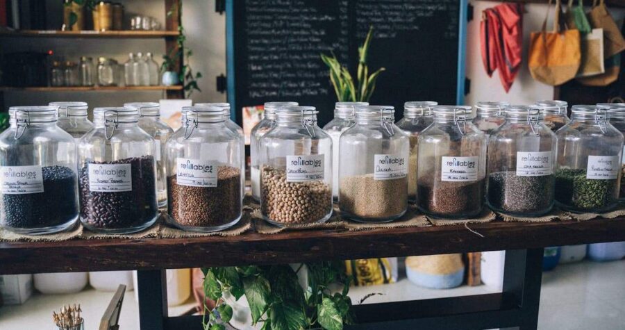 Glass jars with hinged lids containing various seeds, beans, and grains displayed on wooden table in sustainable refill store