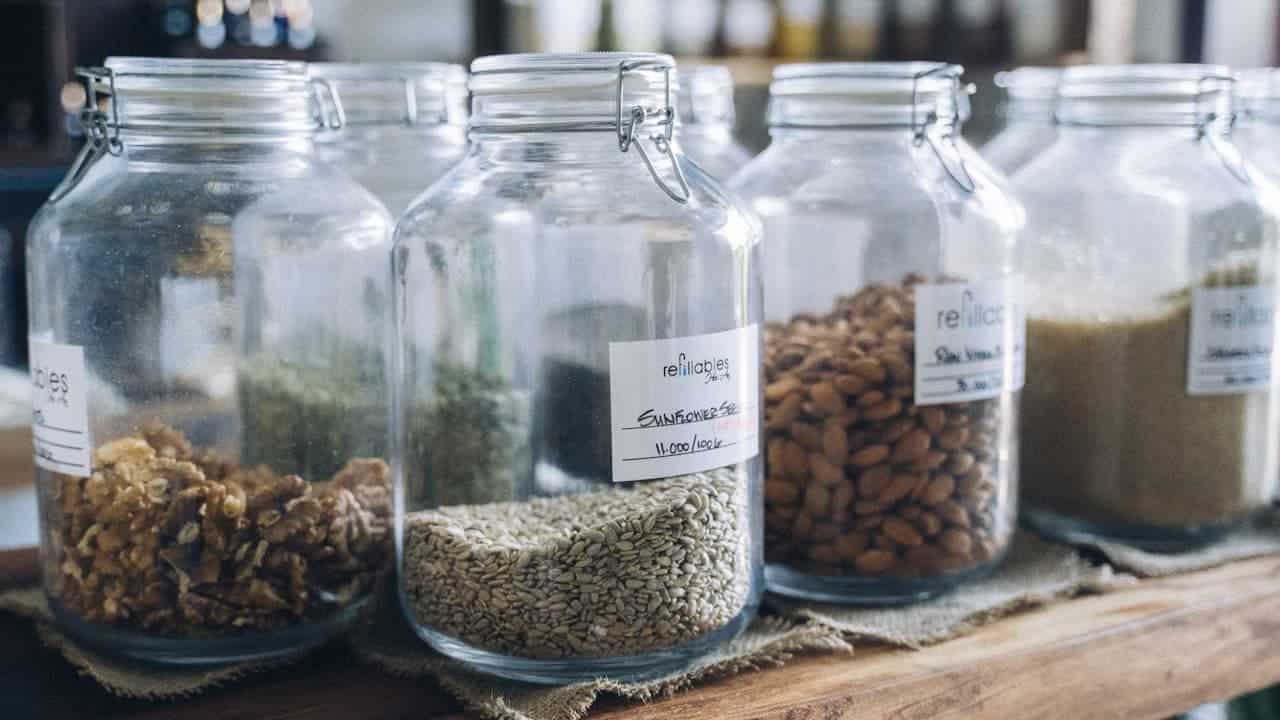 Glass storage jars with hinged lids containing various seeds and grains, labeled and arranged on wooden surface