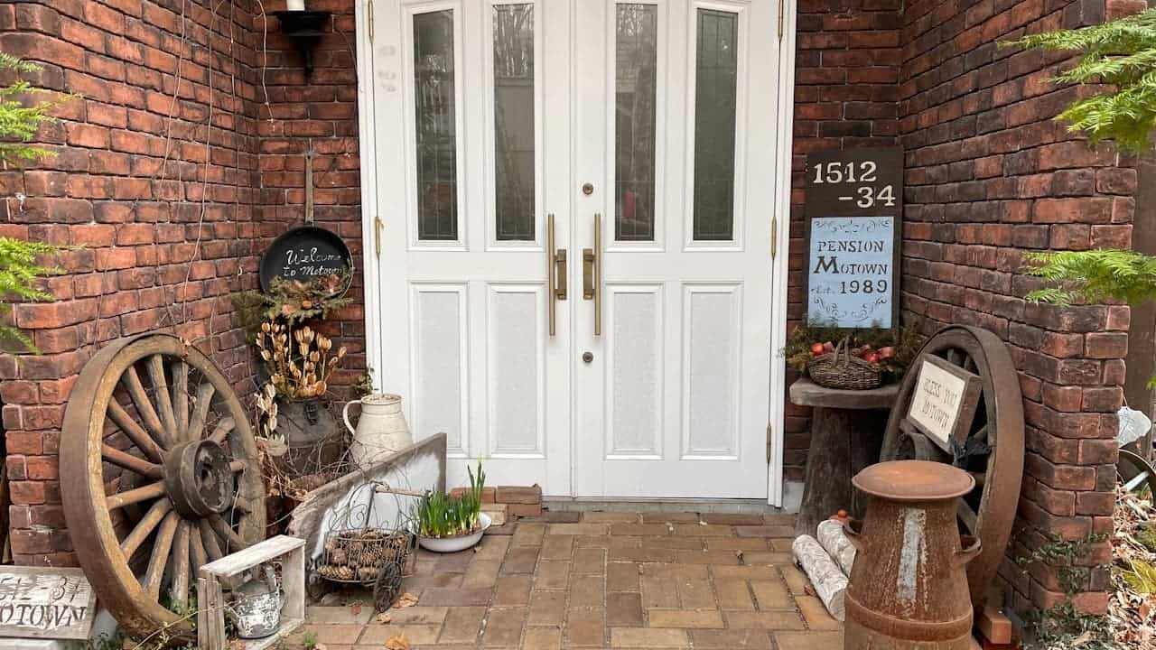 Rustic brick house entrance with white double doors, vintage wagon wheels, pottery, and decorative items creating a charming country-style front porch