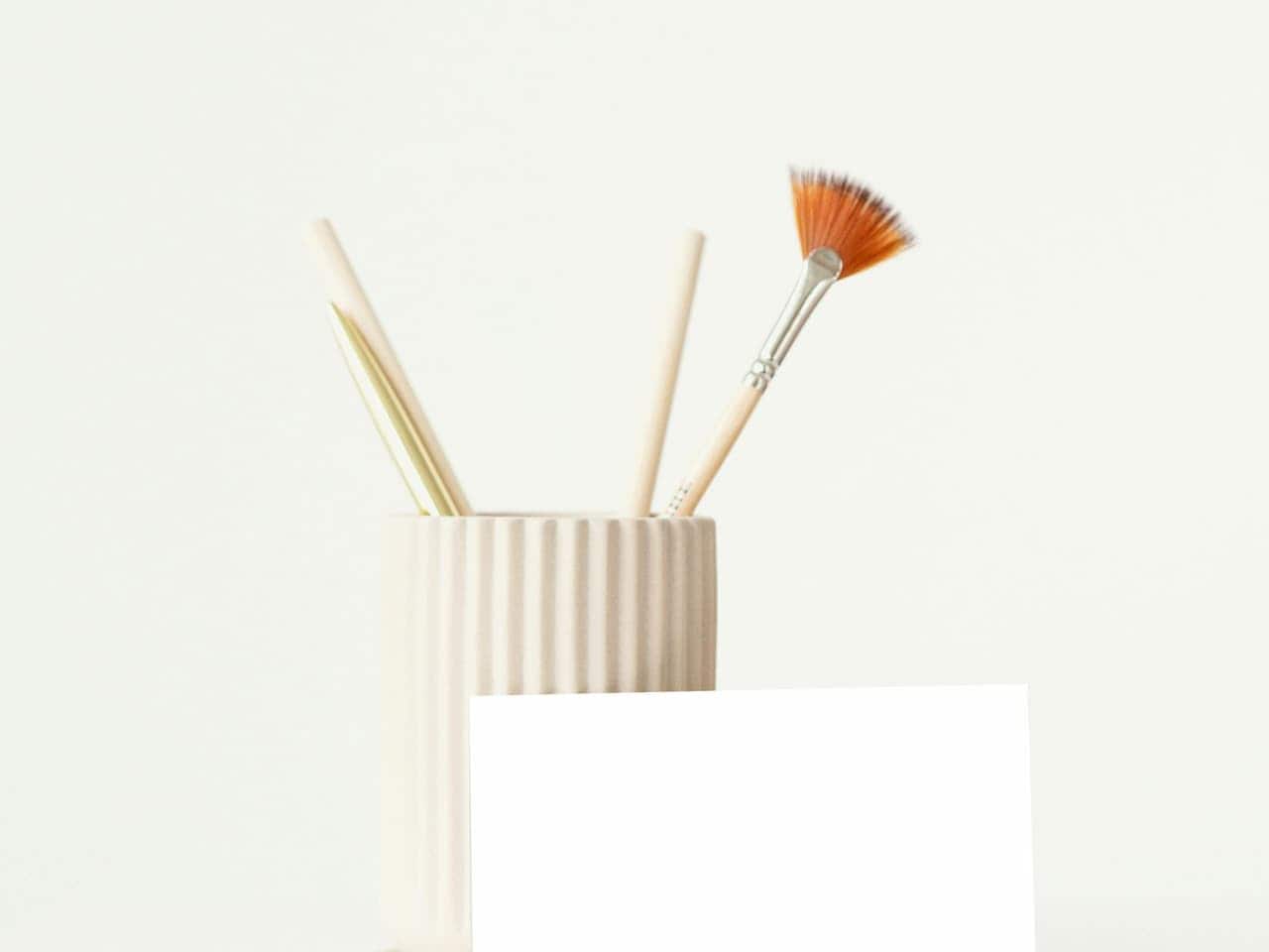 A beige ceramic holder with vertical ridges containing a fan paintbrush, a pencil, and a metallic pen, set against a plain white background