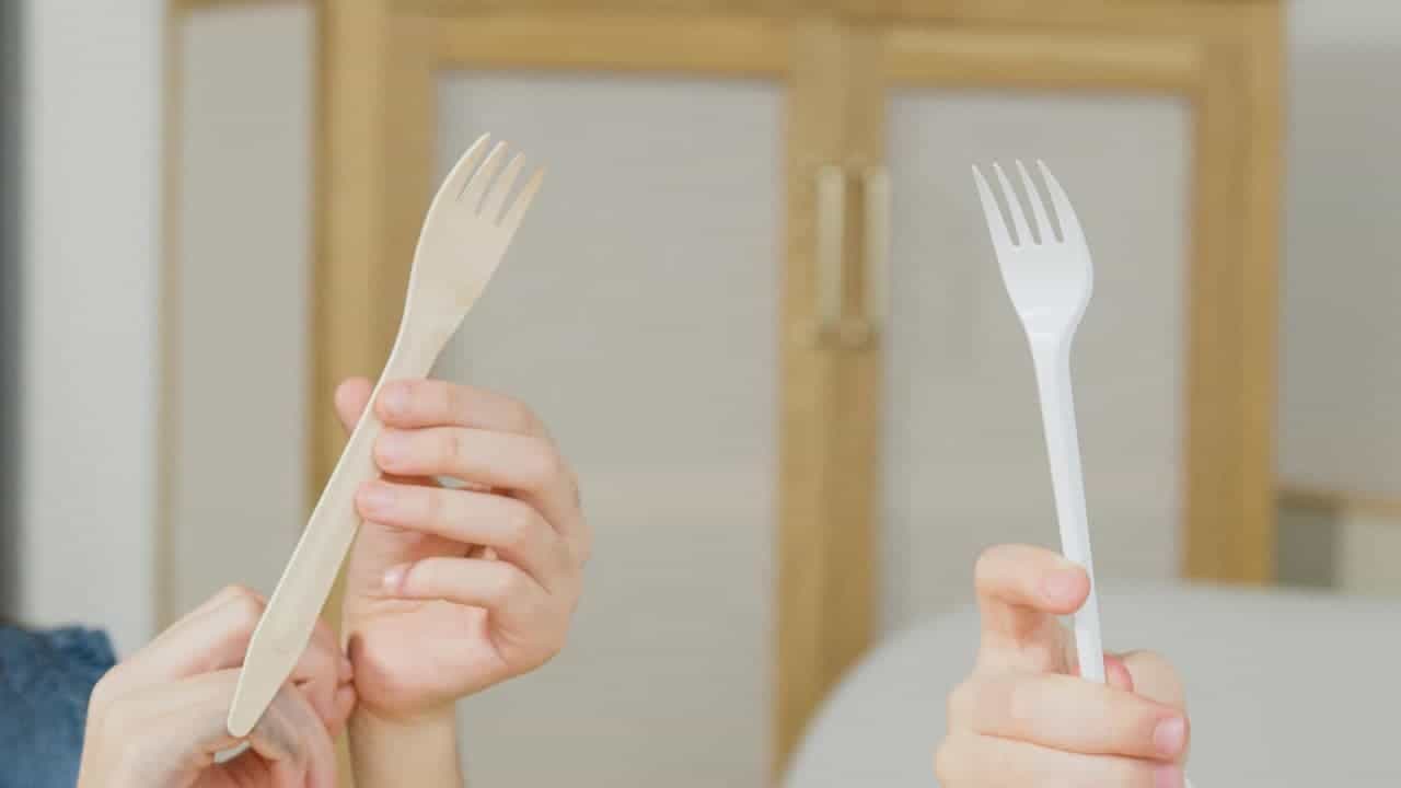 Two hands holding plastic forks, one beige-colored plastic fork, one white plastic fork, indoor setting, light wooden cabinet in background