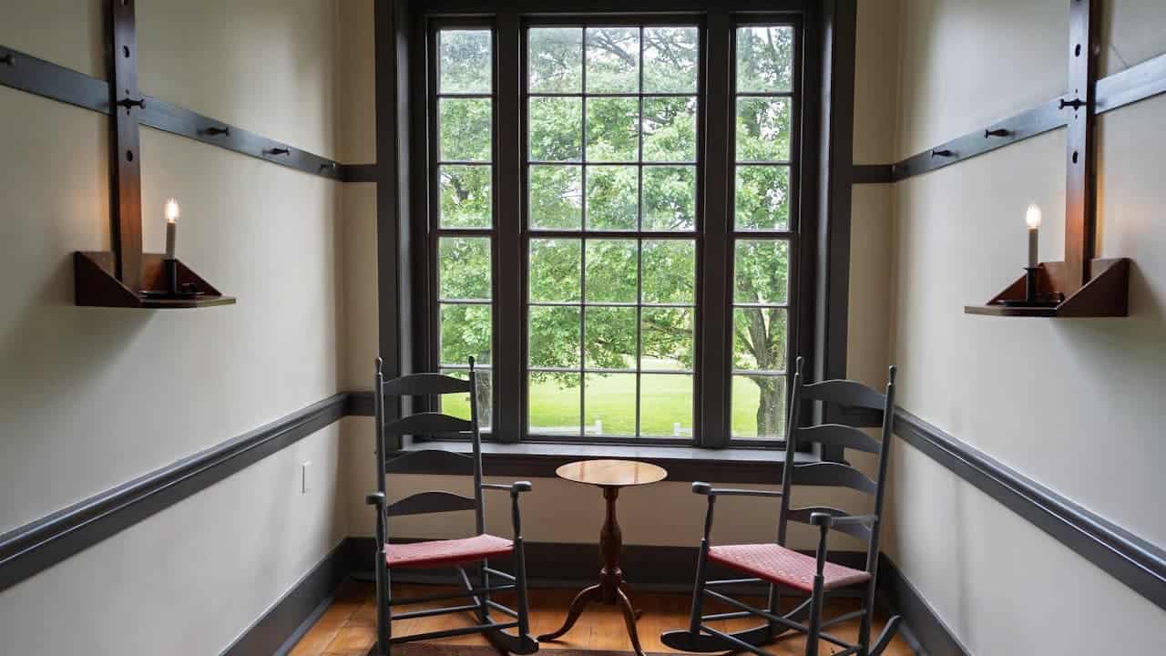 Two black Shaker maple rocking chairs with red cushions positioned by large windows, with candle sconces and small wooden table