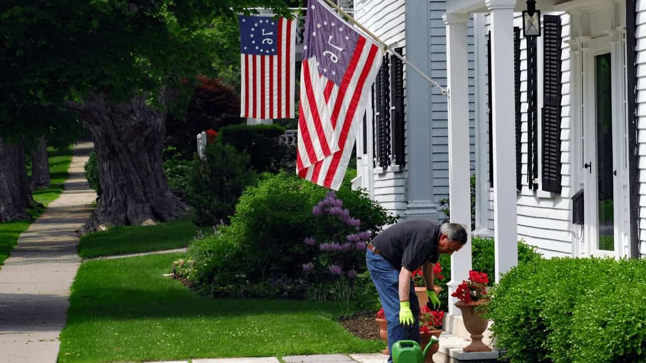 Man gardening in front of white colonial house with American flags, tending to red flowers in planters on porch