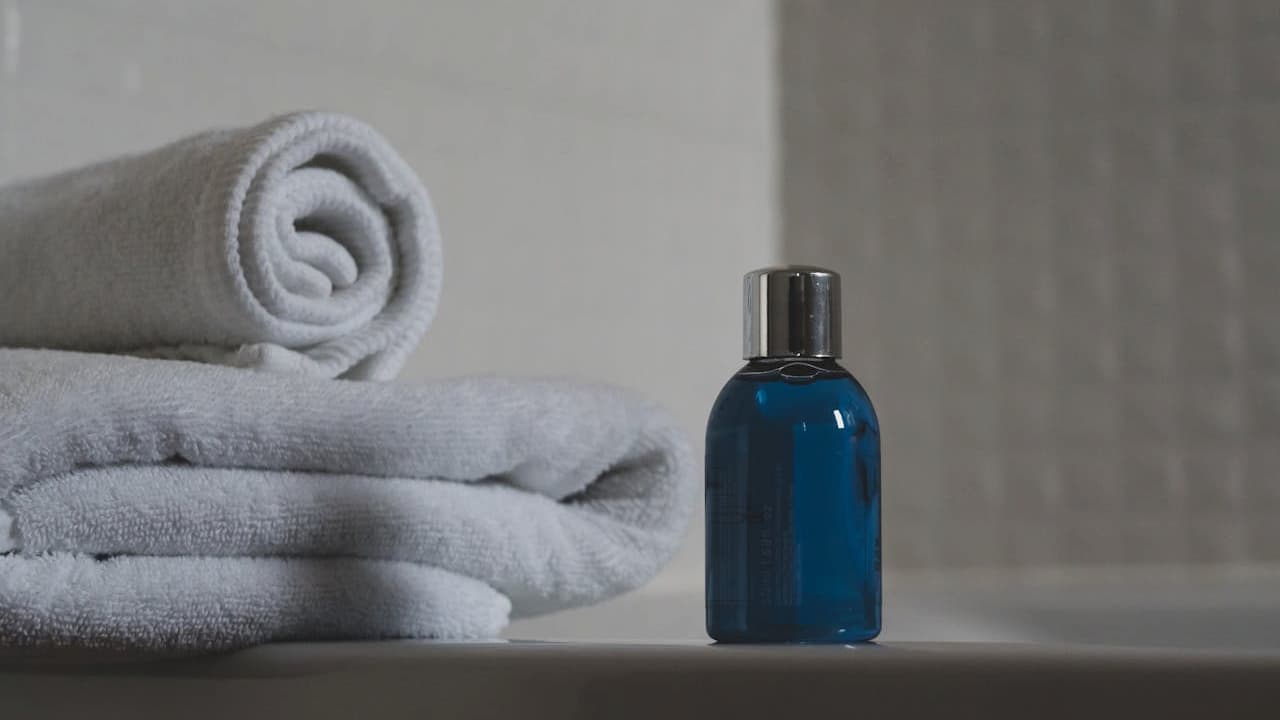 Blue glass bottle with silver cap sitting next to stack of neatly folded white towels on bathroom counter