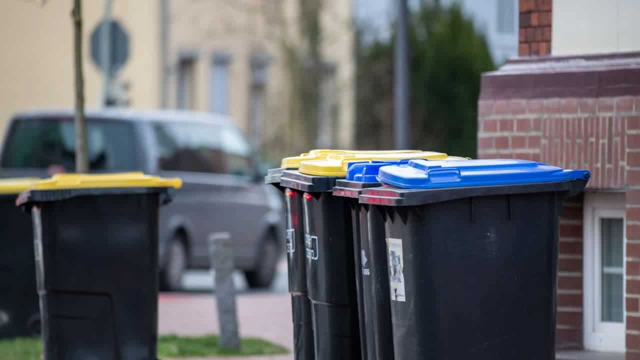 Black recycling bins with yellow and blue lids lined up on residential street with brick houses and car visible
