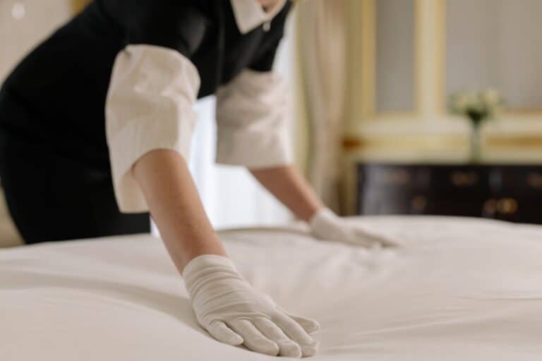 Person in black uniform and white gloves making bed with white linens in hotel or professional cleaning setting