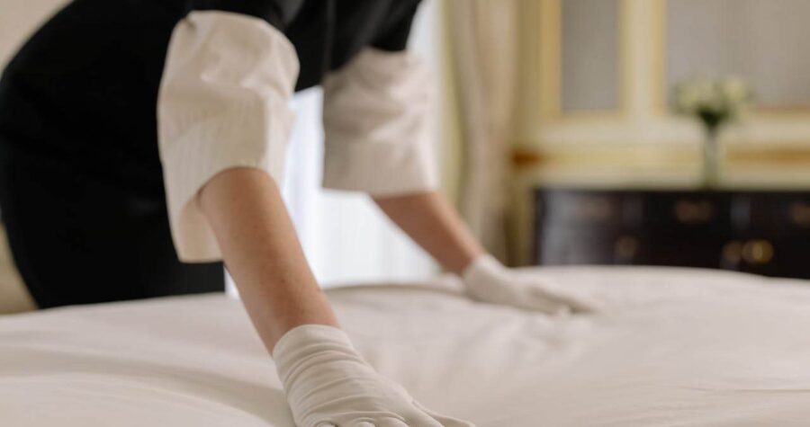 Person in black uniform and white gloves making bed with white linens in hotel or professional cleaning setting