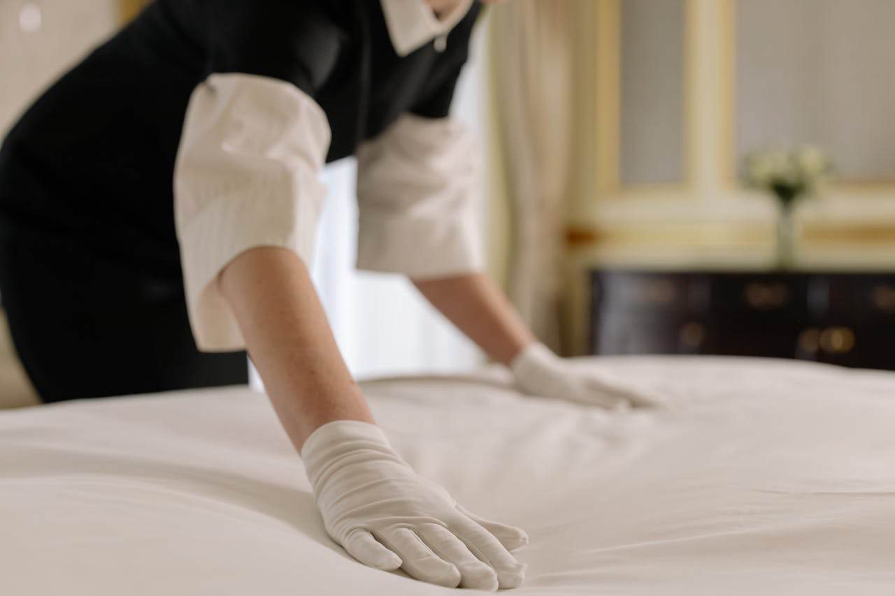 Person in black uniform and white gloves making bed with white linens in hotel or professional cleaning setting