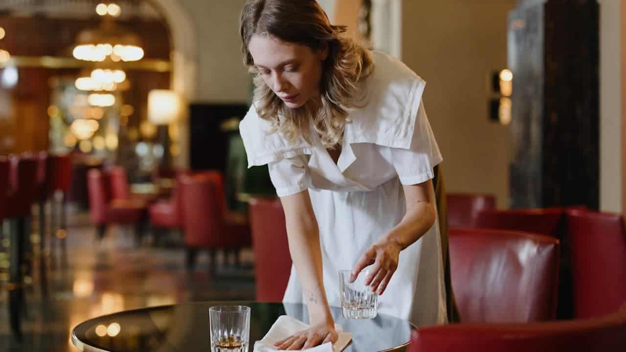 Woman in white uniform cleaning table with cloth in upscale restaurant or hotel lobby with red leather seating