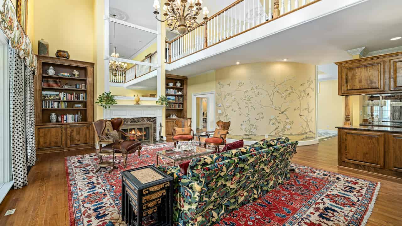 Spacious traditional living room with high ceilings, patterned red Persian rug, floral sofa, classic wood furniture, built-in bookshelves, a fireplace, and a balcony overlooking from the upper floor