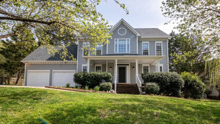 Two-story gray colonial house with white trim, covered front porch, attached garage, and sloped front lawn with mature trees