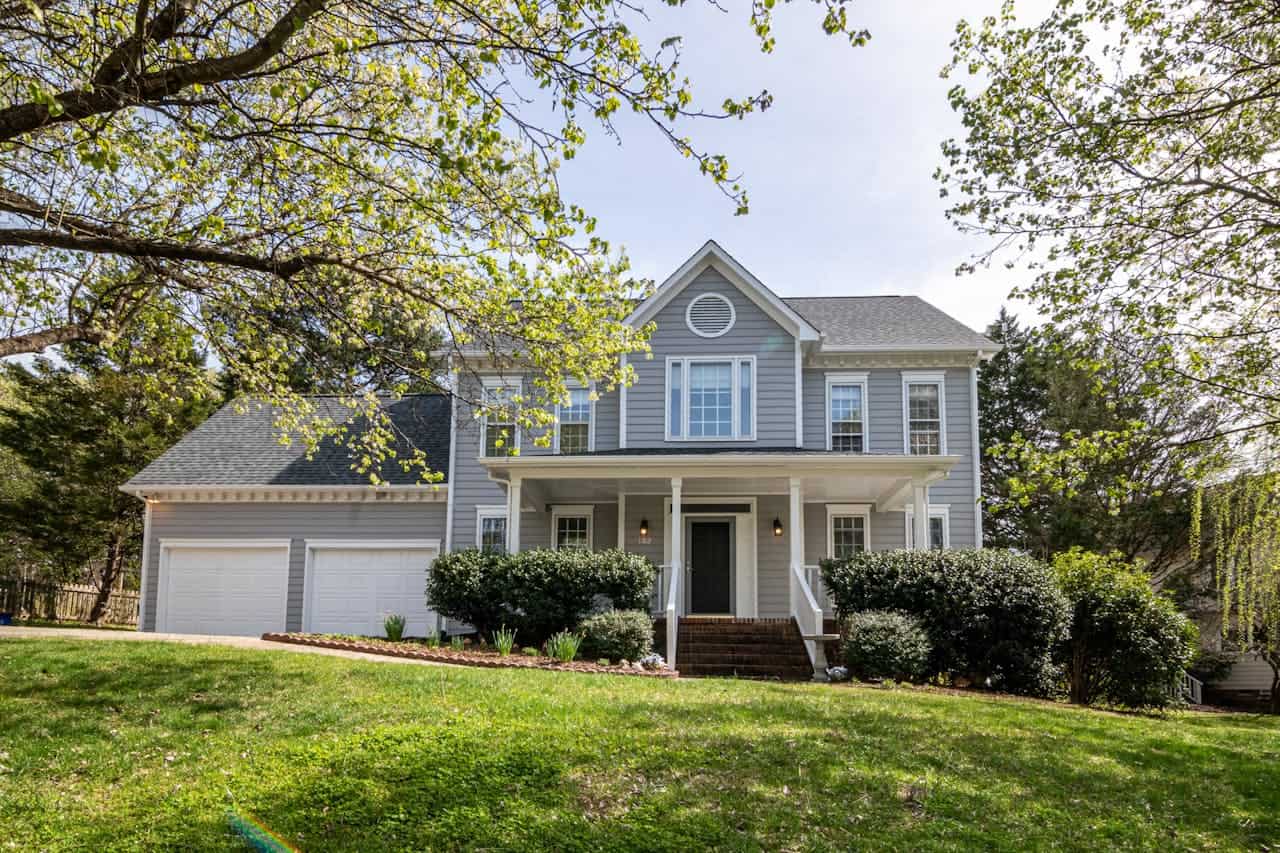 Two-story gray colonial house with white trim, covered front porch, attached garage, and sloped front lawn with mature trees