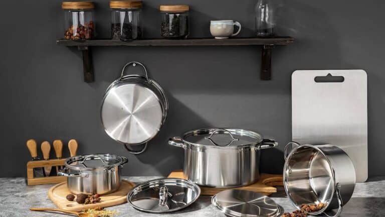 Stainless steel cookware set with pots, pans, and lids displayed on marble counter with wooden utensils and spice jars