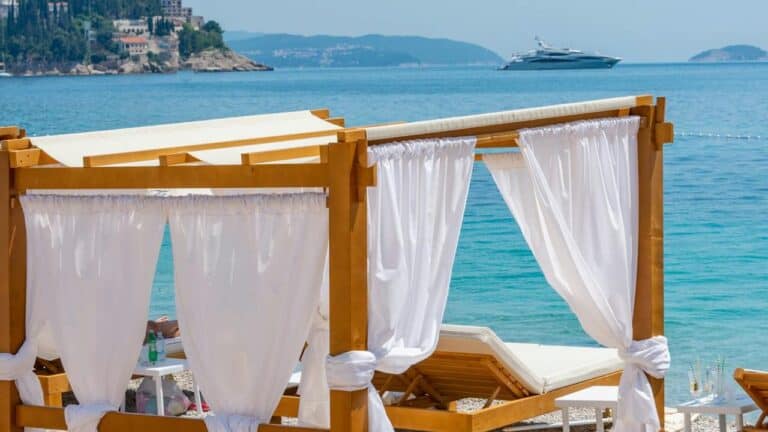 Wooden cabana with white curtains and lounge chairs on a pebble beach, overlooking turquoise sea with a distant yacht