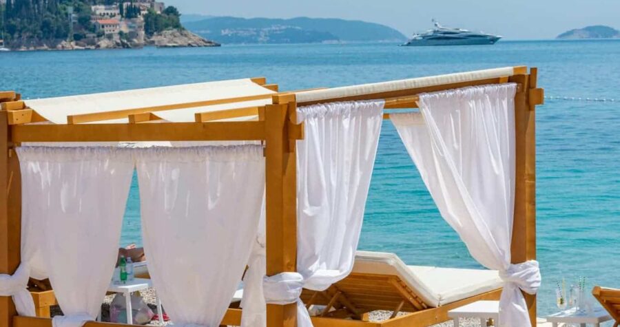 Wooden cabana with white curtains and lounge chairs on a pebble beach, overlooking turquoise sea with a distant yacht