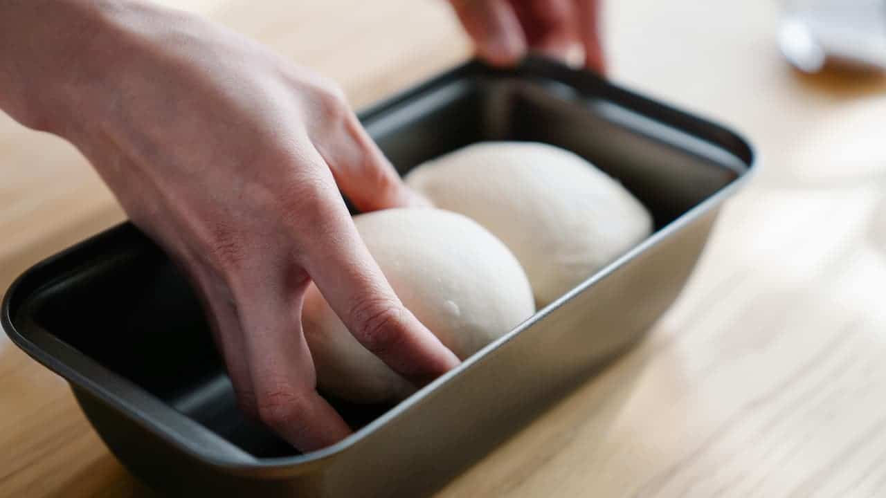 Hands placing two smooth dough balls into a black baking pan