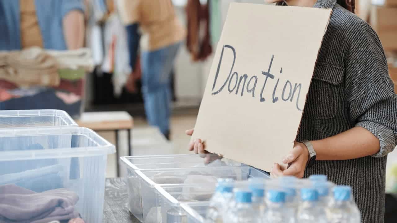 Person in gray sweater holding handwritten "Donation" sign at donation center with clear plastic storage bins and bottles