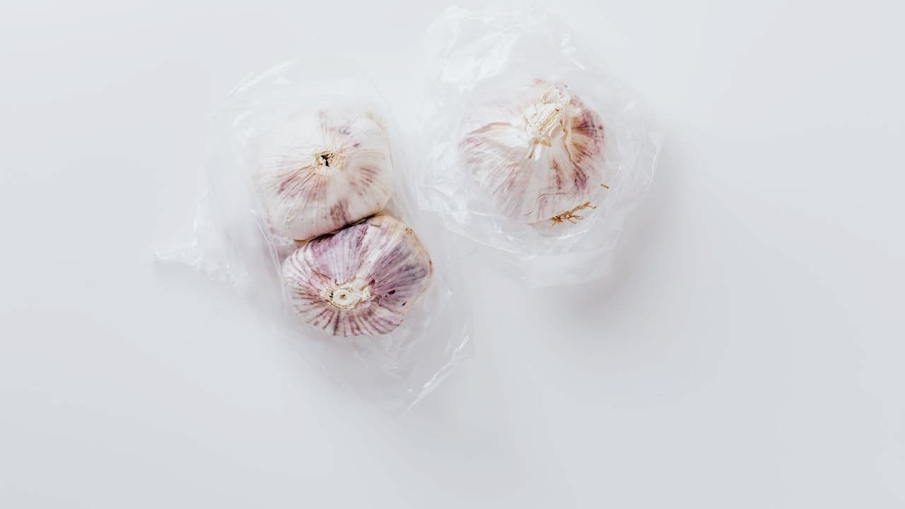 Three whole garlic bulbs with purple-striped papery skin stored in clear plastic bags on white surface