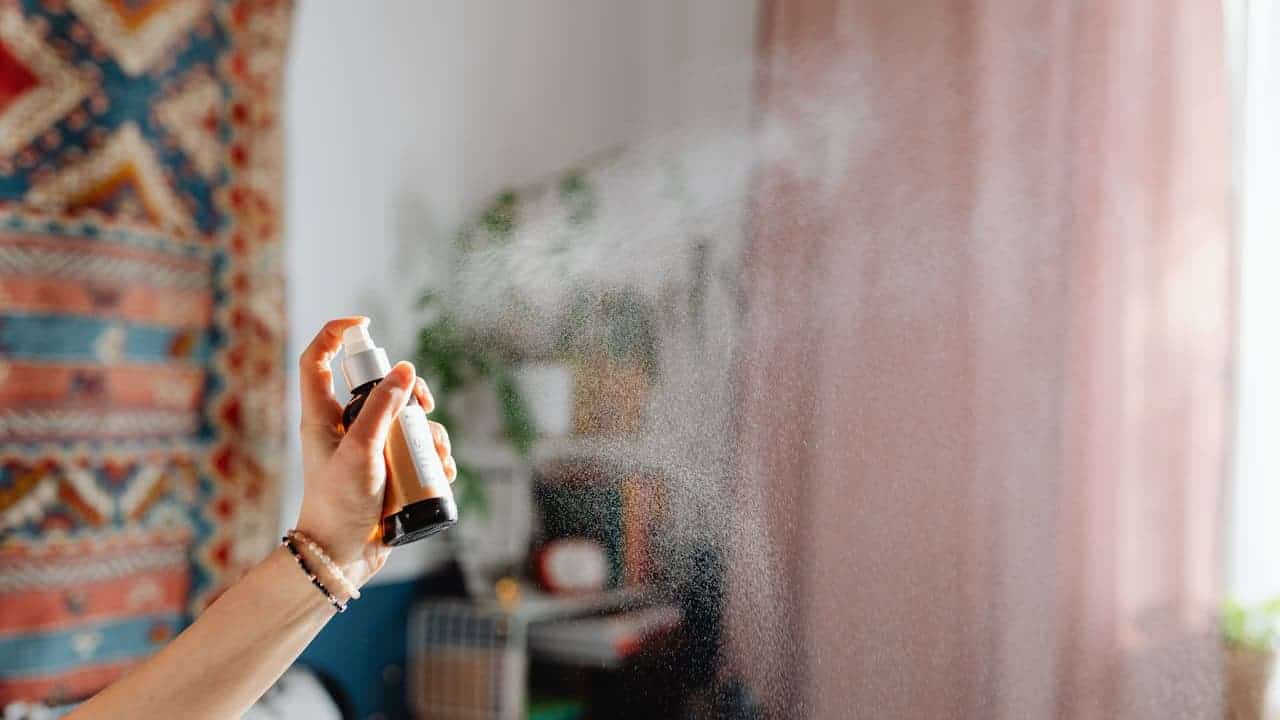 Hand holding spray bottle dispensing fine mist into air, with colorful patterned fabric and blurred home interior background