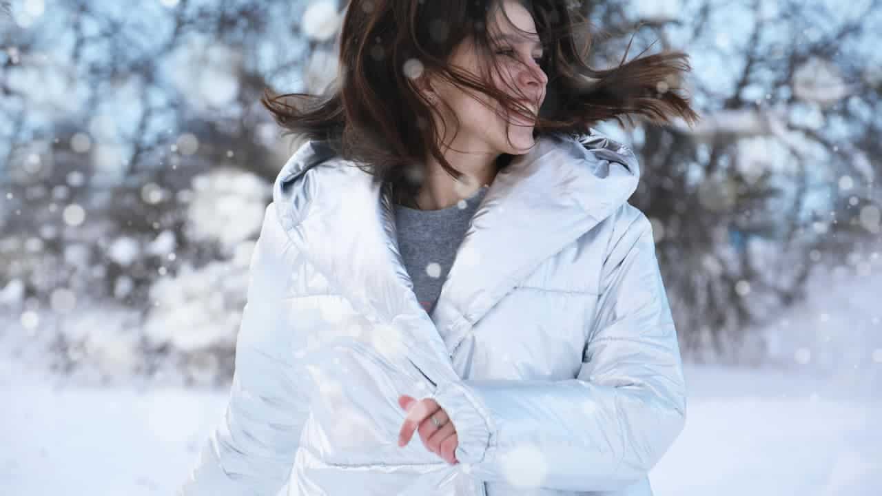 Smiling woman wearing a shiny white winter jacket, standing outdoors in a snowy landscape, with snowflakes falling and wind blowing her hair