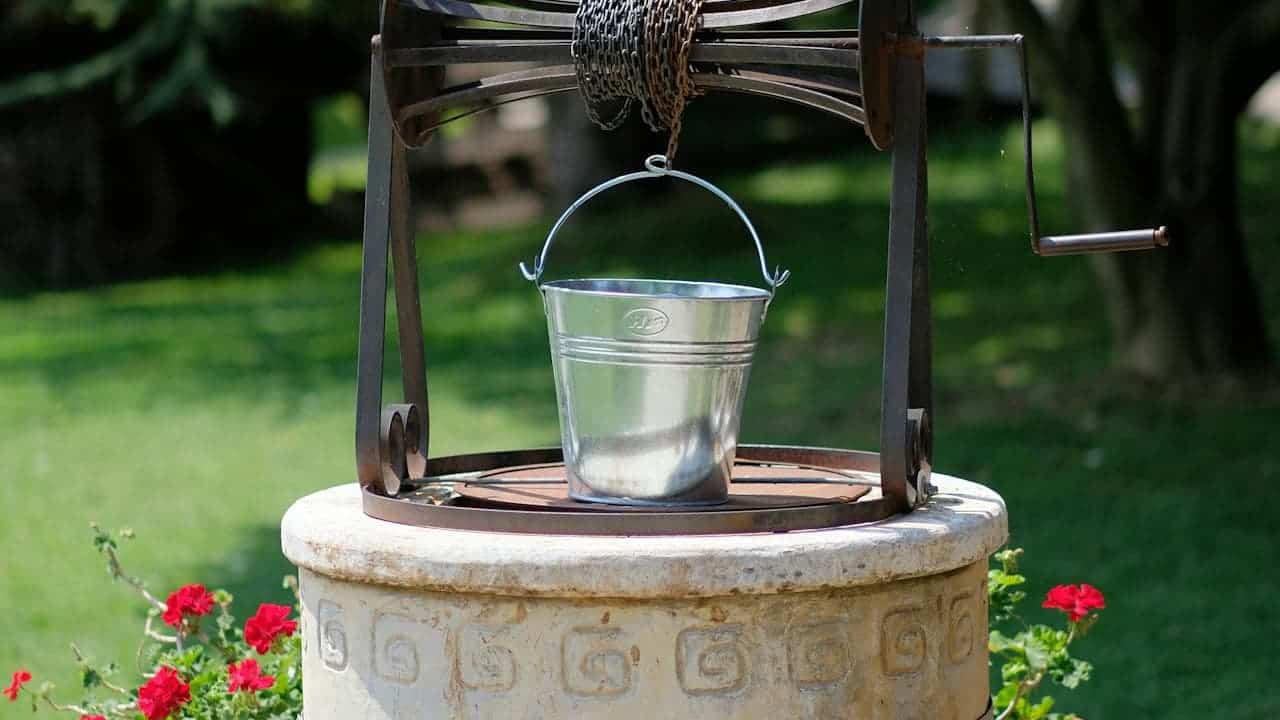 Concrete wishing well with a bright metallic bucket hanging at the center, set in an outdoor garden or yard