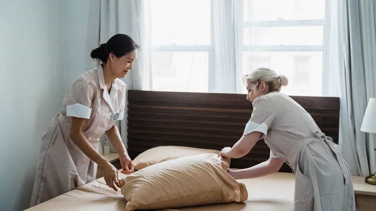 Two hotel housekeepers in uniforms making bed together, one in white uniform, one in beige, bright hotel room
