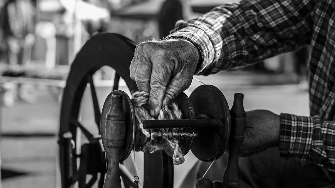 Elderly hands in plaid shirt cleaning vintage black spinning wheel mechanism, demonstrating traditional fiber craft in workshop setting