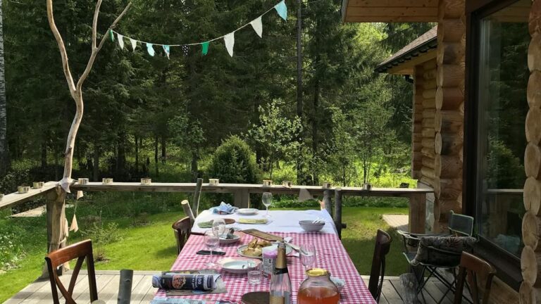 Outdoor dining setup on wooden deck with red gingham tablecloth under log cabin porch, surrounded by forest and bunting