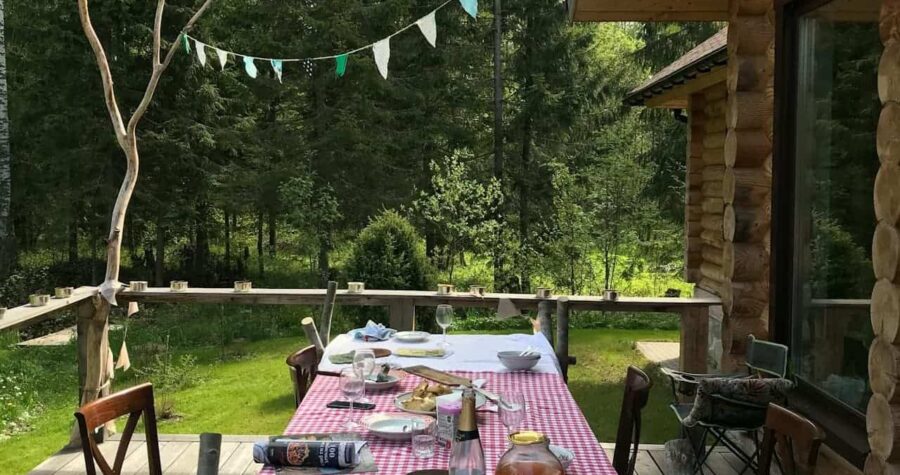 Outdoor dining setup on wooden deck with red gingham tablecloth under log cabin porch, surrounded by forest and bunting