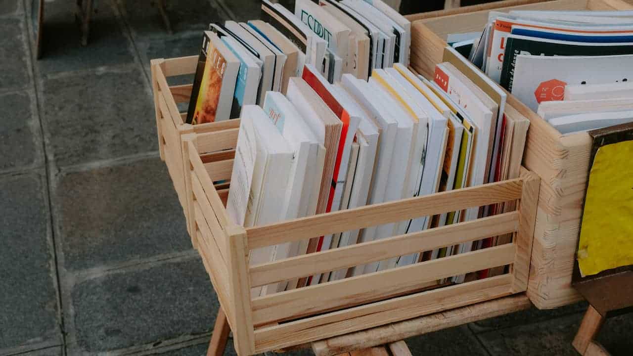 Wooden crates filled with assorted books and magazines, arranged upright, placed on wooden stands over a tiled pavement