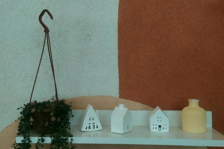 White wall shelf with small decorative house ornaments, a yellow vase, and a hanging potted plant against a textured wall with abstract curves in beige