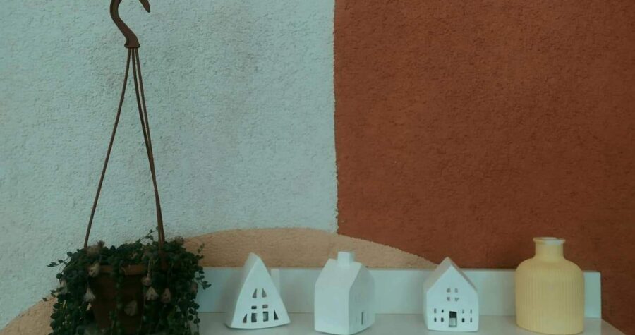 White wall shelf with small decorative house ornaments, a yellow vase, and a hanging potted plant against a textured wall with abstract curves in beige
