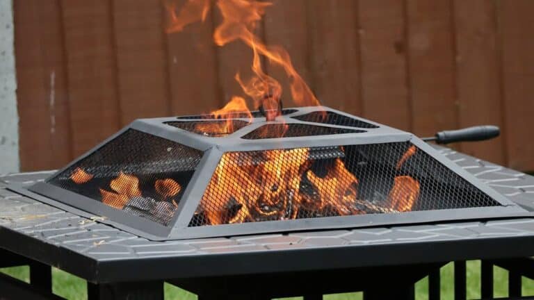 Square metal fire pit with mesh screen and burning wood logs producing orange flames on outdoor table