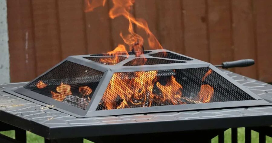 Square metal fire pit with mesh screen and burning wood logs producing orange flames on outdoor table
