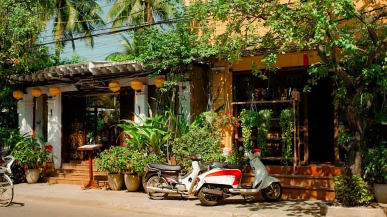 Traditional Asian shophouse with mismatched architectural elements, motorbikes parked outside, surrounded by lush tropical plants and hanging baskets