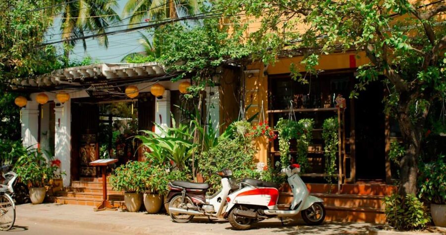 Traditional Asian shophouse with mismatched architectural elements, motorbikes parked outside, surrounded by lush tropical plants and hanging baskets