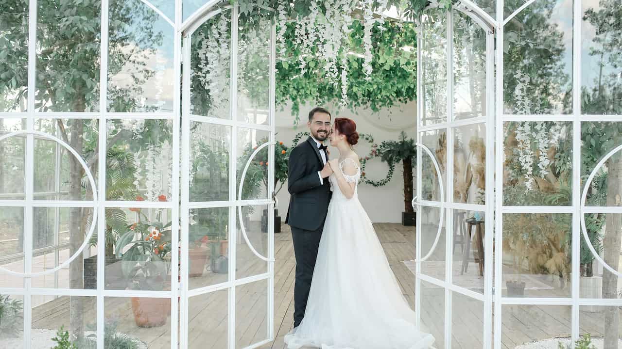 Newlywed couple standing in front of a glass greenhouse, dressed in wedding attire and sharing a joyful moment