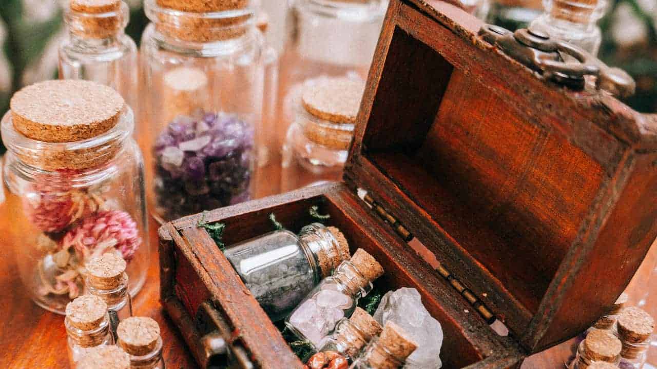 A small vintage-style wooden chest sits open on a wooden surface, revealing an assortment of miniature glass bottles sealed with corks